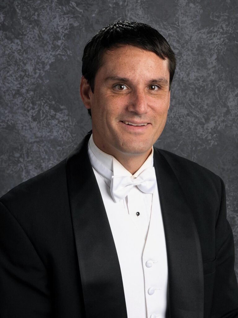 Paul Kile, Minnesota Symphonic Winds associate conductor, smiles into the camera in this formal portrait wearing a black tuxedo with white shirt, bowtie, and vest.