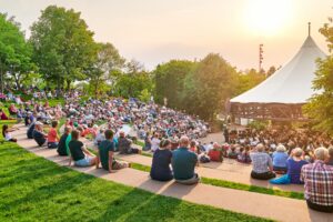 MSW performs outdoors under a pavilion. The summer sun is setting in the background as audience members enjoy the performance.