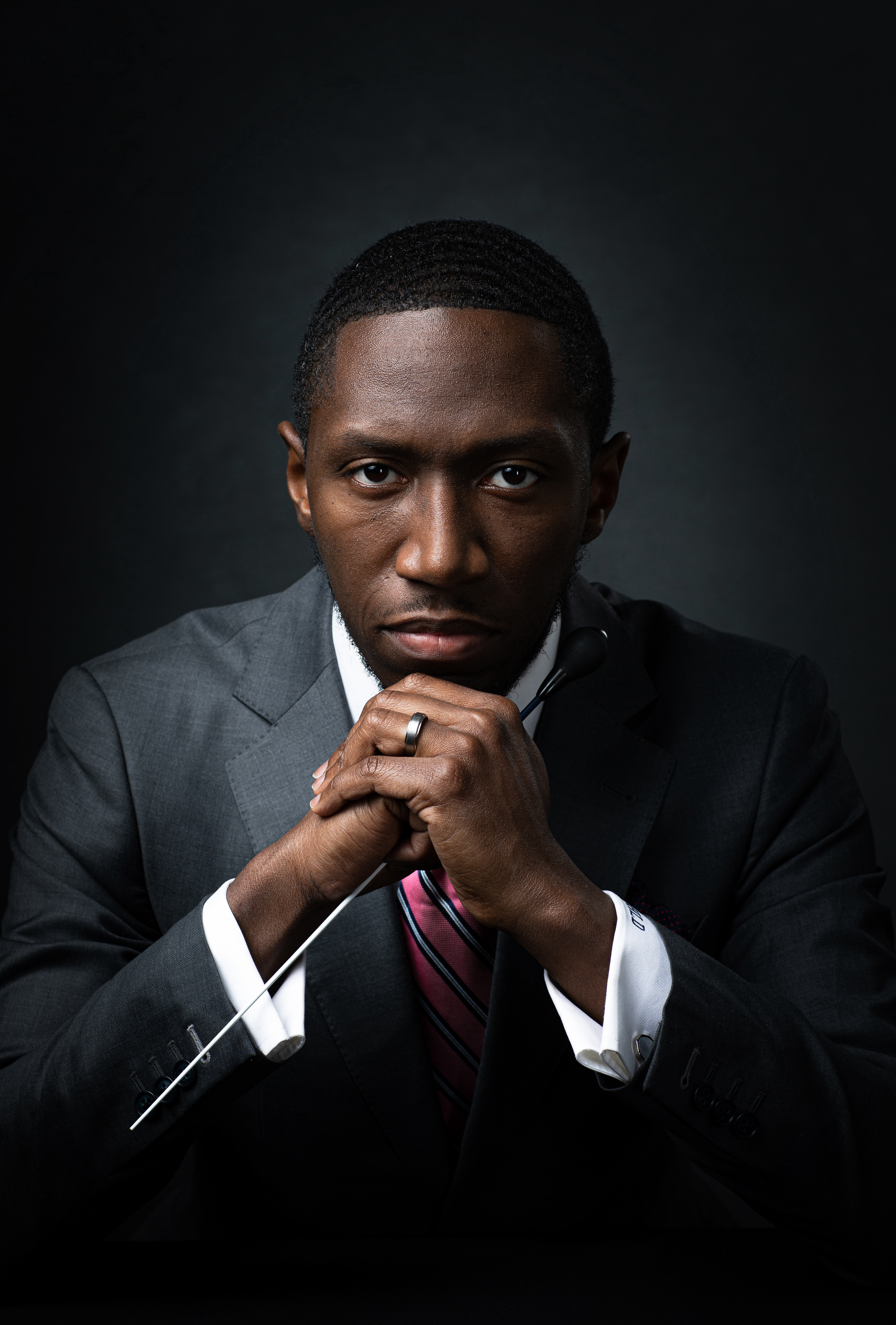 Dr. Henry Dorn looks intensely at the viewer. He is wearing a grey suit and has his hands folded by his chin. He is holding his baton with the tip facing down towards the left along his arm.