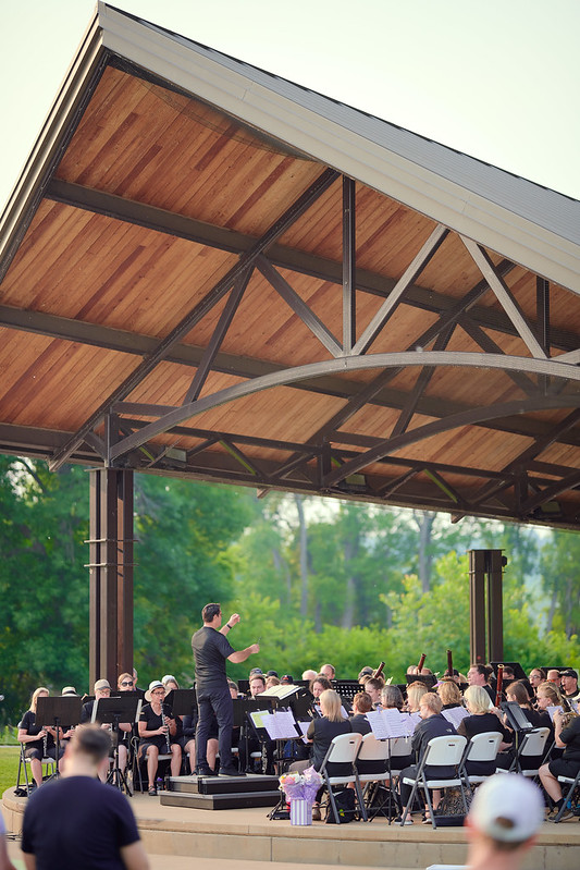 MSW performs on a stage under a tall pavilion in a park. The photograph is taken from the audience perspective, about 45 degrees right from center, looking at the stage. The stage fills the lower quarter of the frame, with a couple audience members in the foreground. A view of the ceiling of the pavilion fills the top half of the frame. The ceiling is paneled in warm strips of wood supported by metal arches curving over the stage. Behind the pavilion lush green trees and a hazy summer sky create a beautiful backdrop.