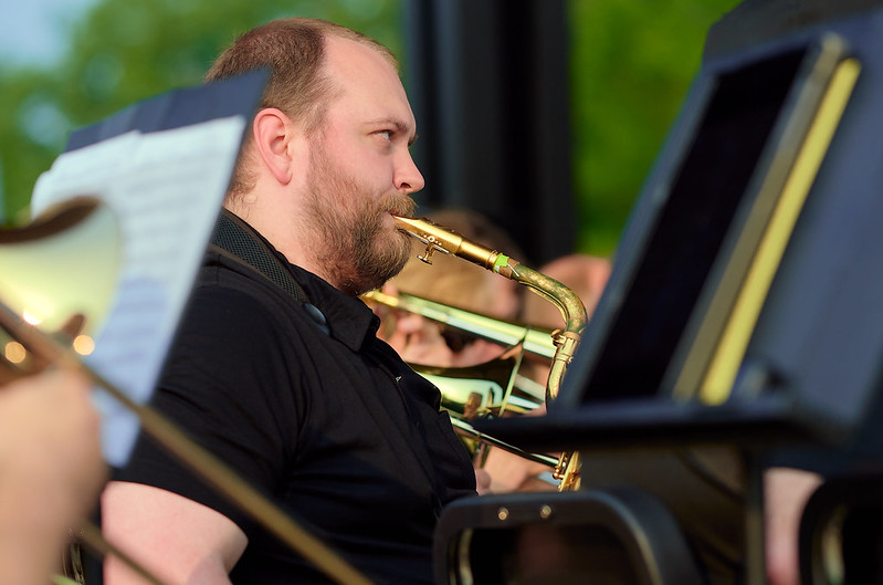 Looking between music stands the viewer sees a bearded male musician playing the baritone saxophone in an outdoor setting. He is wearing concert black attire and only the mouthpiece and very top of his instrument are visible. Other musicians playing trombones sit around him.