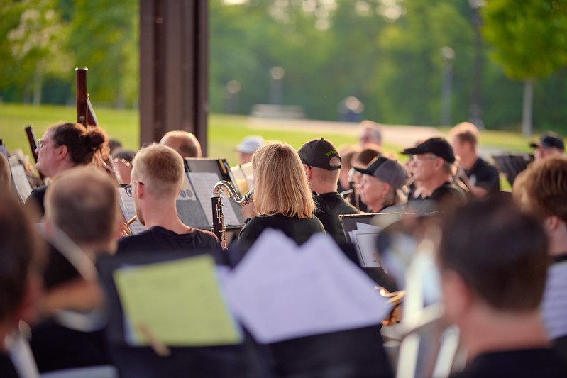 Viewed from the back-lefthand side of the stage, members of MSW perform outdoors under a covered pavilion. Centered in the frame is a lovely woman (both inside and out) playing the bass clarinet. In the background is a grassy lawn with green trees. The light has a special soft quality as a hazy yellow sunset is just beginning.