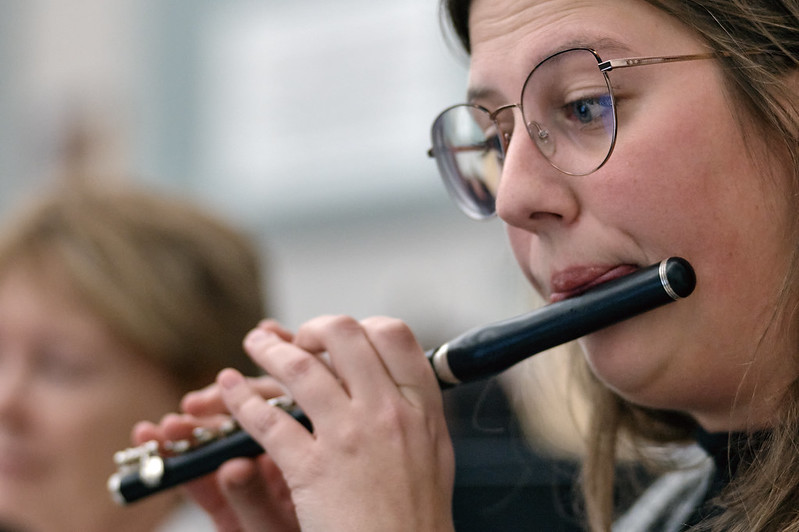 A close up of a woman playing the piccolo. She is on the right side of the frame lookin down and to the left. A flute player is in the background.