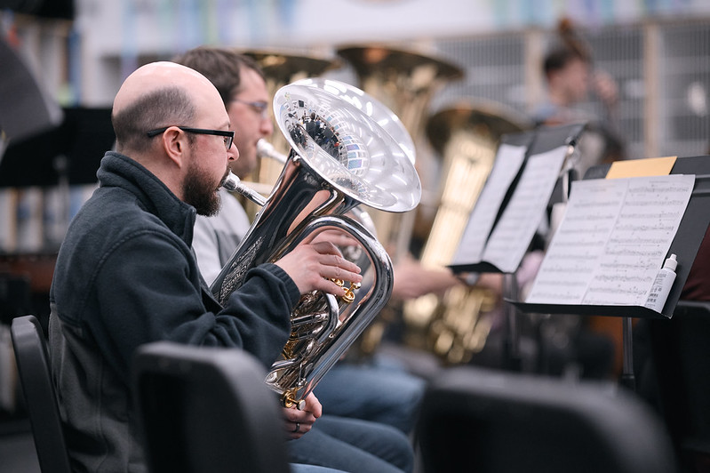 Image of a man in profile playing the euphonium. He is seated on the left edge of the frame facing right. His stand is on the far right edge of the frame. Other euphonium and tuba players are in the background.
