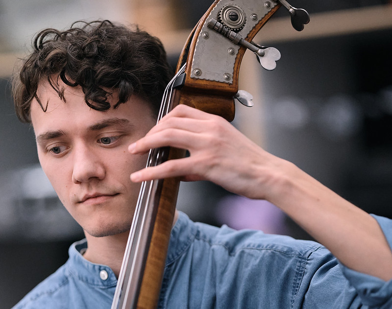 A young man in a blue denim shirt plays the double bass. He is seen from the shoulders up, facing the viewer, with his face along the left of the frame. He looks down towards his music stand out of the frame on the left. His hand presses strings near the top of the instrument near his face. His elbow points out of the frame to the lower right corner.