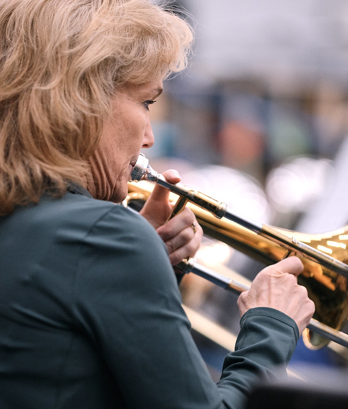 A woman playing the trombone. She is wearing a blue shirt and her face is shown in profile from over her right shoulder.