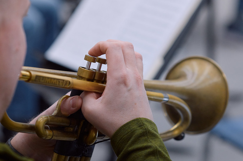 Close up of a musician playing a trumpet. His hands are centered in the frame and only his cheek is visible. He is on the left side of the frame looking away and towards the right.