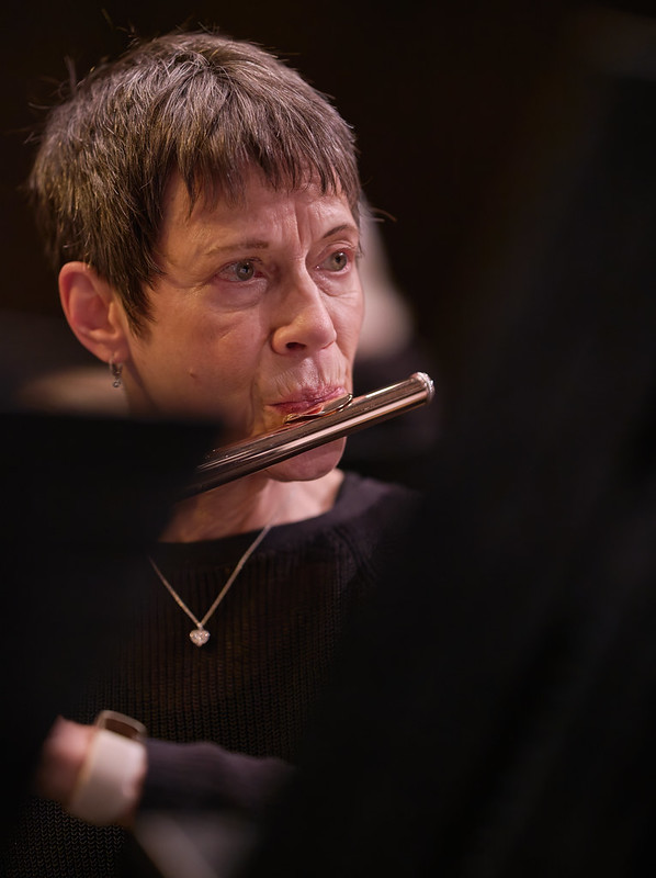 A woman in black concert attire and a delicate necklace plays the flute. She is seen from her shoulders up facing the viewer, and is seated along the left side of the frame looking towards the right. Her instrument and shoulders are partially obstructed from view by music stands.