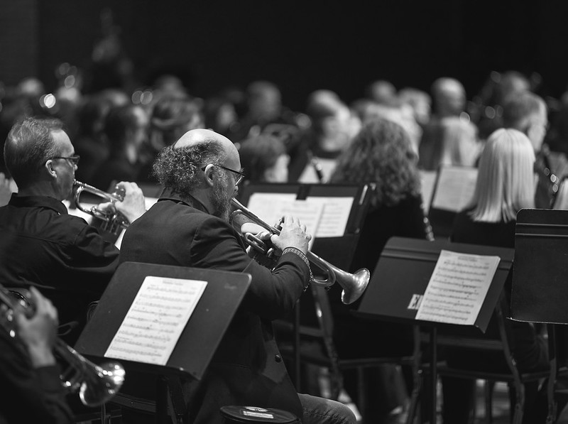 A black and white image of the ensemble playing a concert. Two male trumpet players are featured in the middle left partially looking away from the camera.