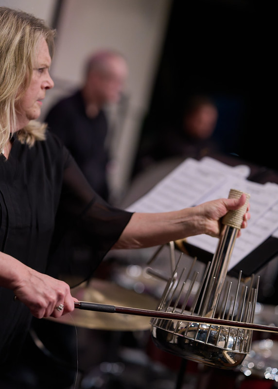 A woman in concert black attire plays the percussion with a bow. She stands on the left side of the frame looking to the right. Other percussionists are seen in the background.
