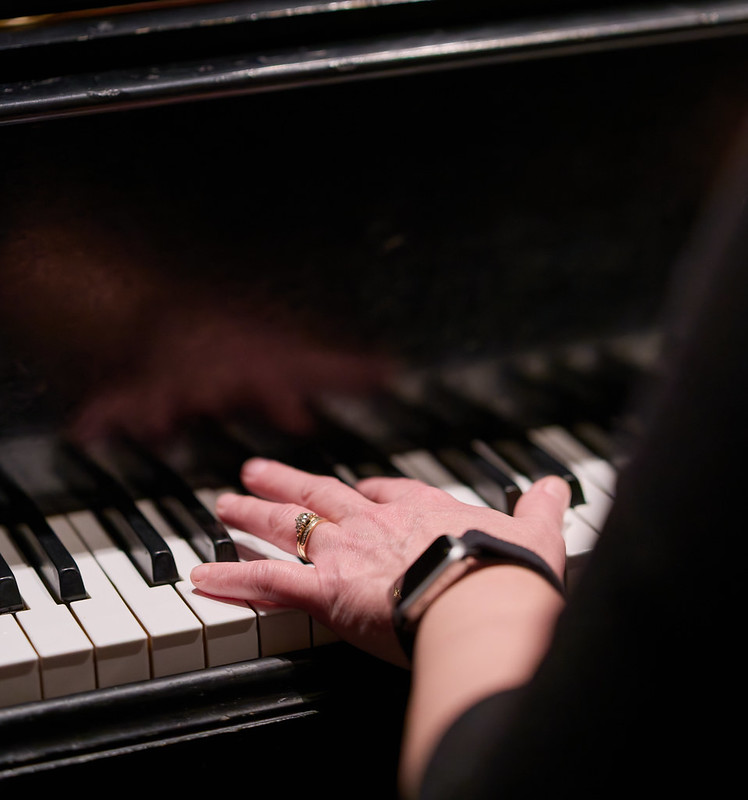 A closeup image looking over a woman's left shoulder while she plays a grand piano. The keys are slightly angled from the middle right of the frame to the bottom left. the viewer sees only the outer edge of her left arm and her left hand. She wears concert black attire, a wedding ring, and a watch.