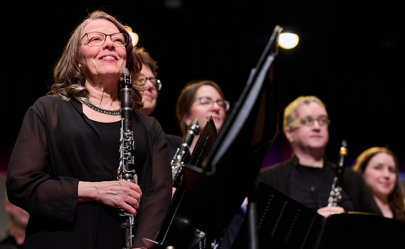 An image of four smiling clarinet players standing to acknowledge the audience after performing a piece. They are wearing black concert attire.
