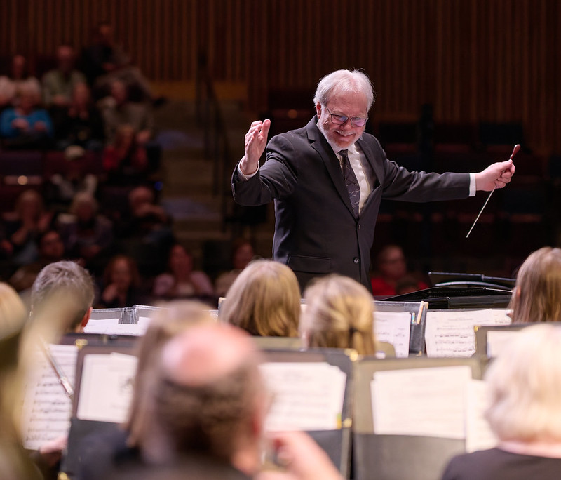 An image of our conductor, Dr. Timothy Mahr, acknowledging the band during a performance with the ensemble. He is on the podium facing the band. He is dressed in a black suit, is smiling, and his arms are stretched wide. One hand holds his baton pointing downwards. Several musicians and stands are visible in the foreground.