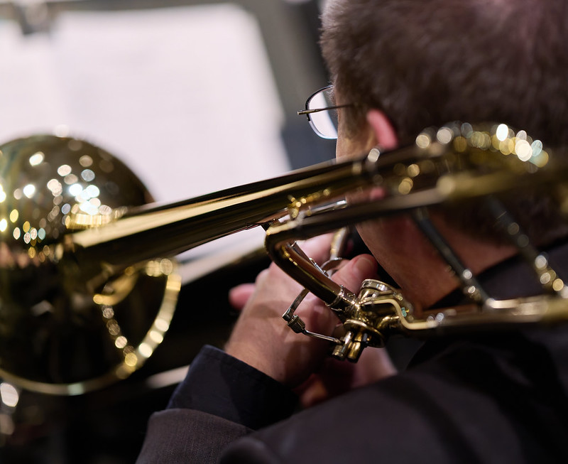 A man wearing concert attire plays the trombone. He is on the right side of the frame looking away from the viewer towards the left. the left side of this head, his shoulder, and left hand are visible. His trombone fills the bottom of the frame from right to left over his left shoulder, with the bell furthest away. His music stand is in the background. 
